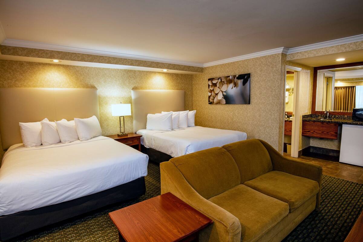 Habitación de hotel con dos camas, un sofá, una mesita y un baño visible a través de una puerta abierta.