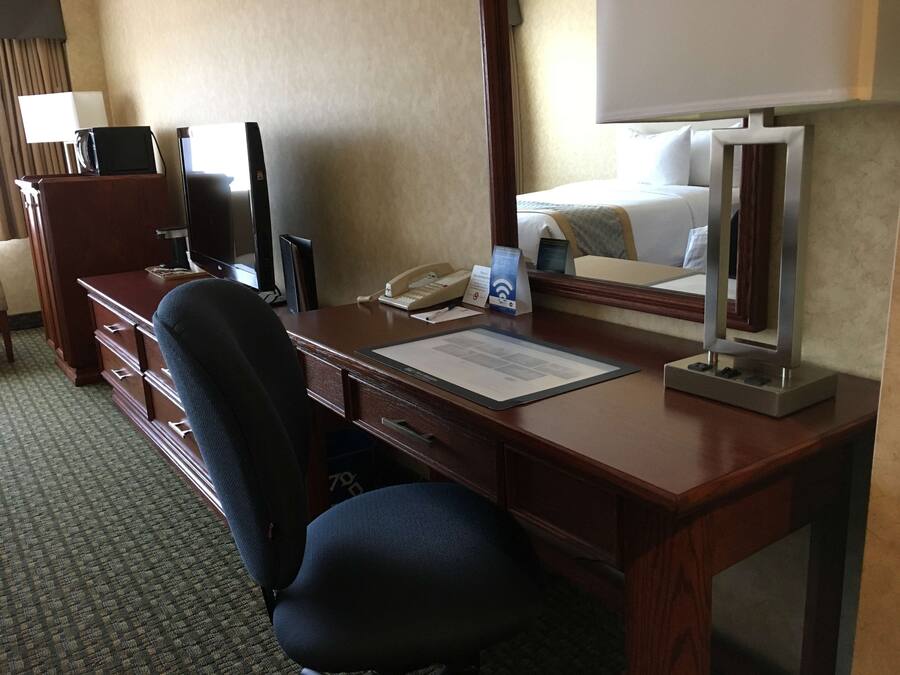 Une chambre d’hôtel avec un bureau en bois, une chaise, un téléphone, un miroir, une télévision et un lit recouvert d’une couette blanche.