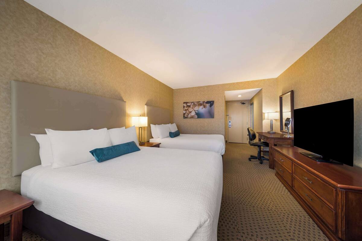 Habitación de hotel con dos camas, un escritorio, un televisor y un baño visible a través de una puerta abierta.
