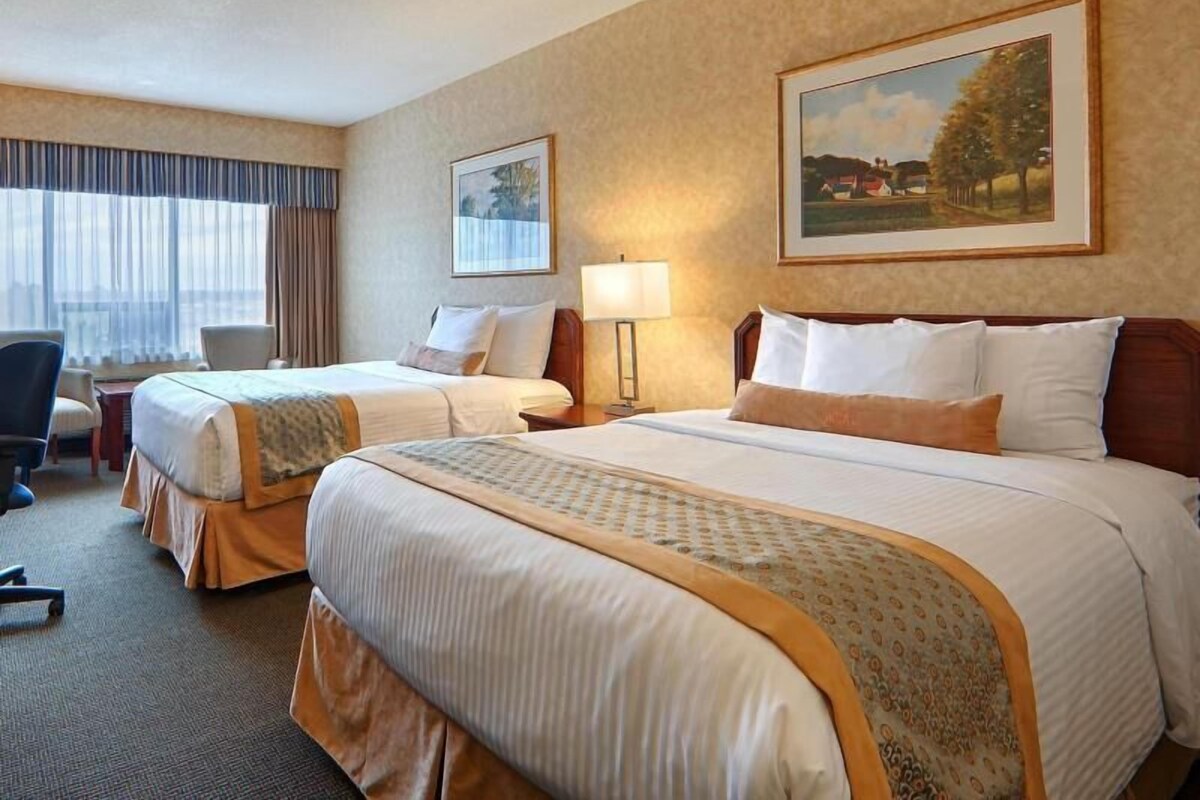 A hotel room with two beds, a desk, a chair, and two paintings on the walls.