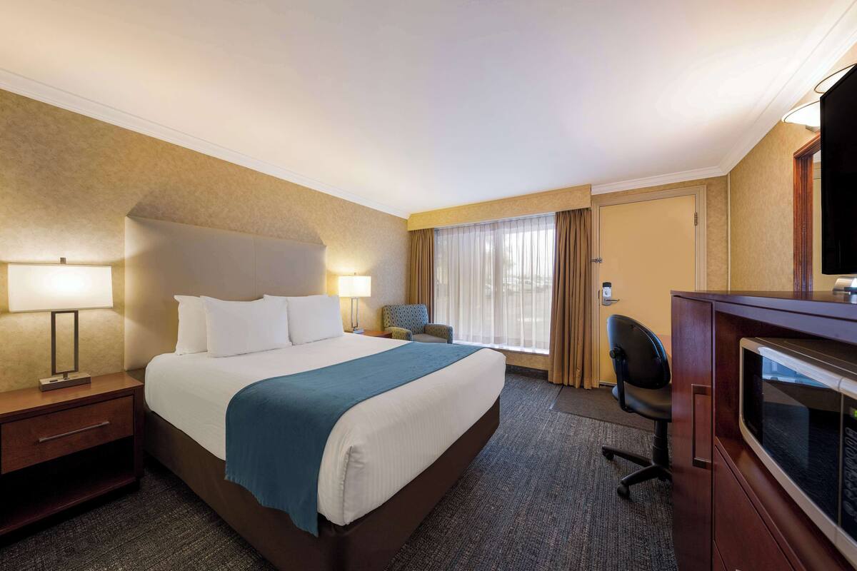 Habitación de hotel con una cama grande, dos mesitas de noche con lámparas, un escritorio con una silla, un televisor y una ventana con cortinas.