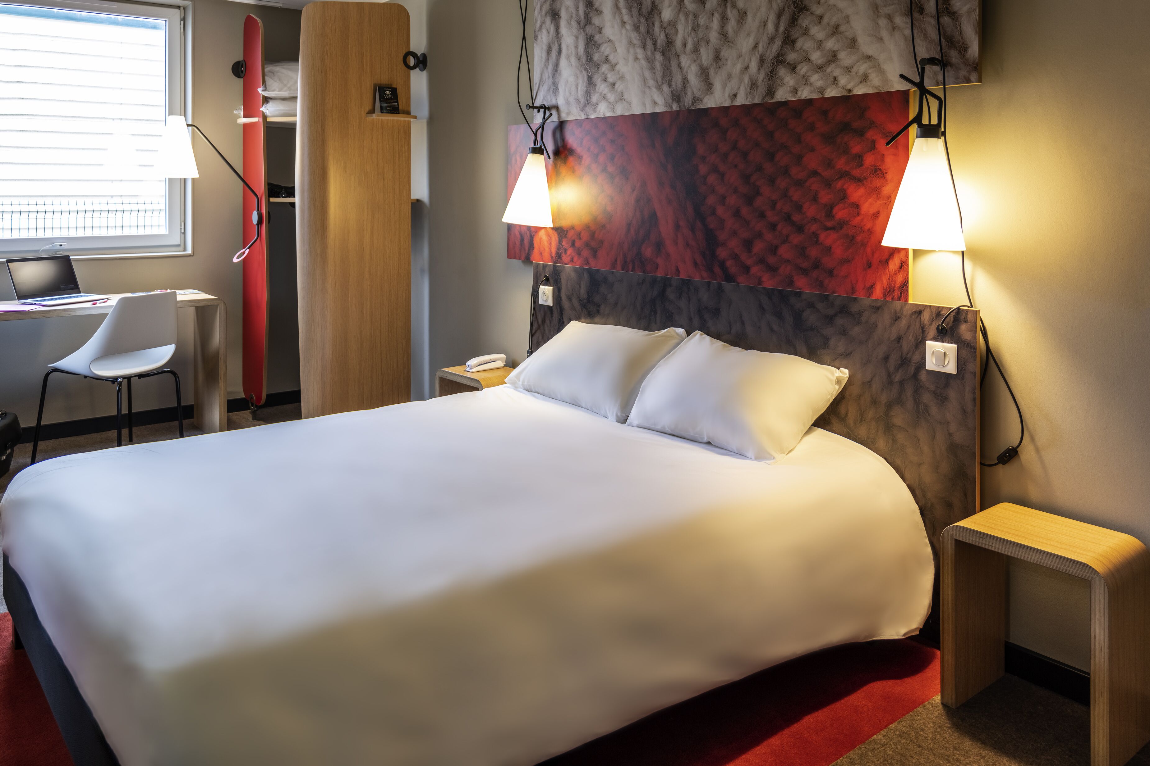 A hotel room with a large bed, a desk with a chair, a red surfboard, and two wall-mounted lamps.
