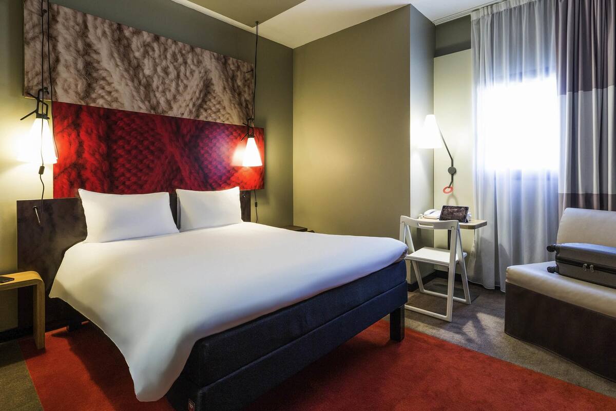 A hotel room with a large bed, bedside lamps, a desk with a chair, and a window with curtains.