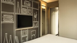 A hotel room with a large flat-screen TV mounted on the wall, a bed, and a door leading to a bathroom. - ibis Bordeaux Centre Gare Saint Jean (Bordeaux)
