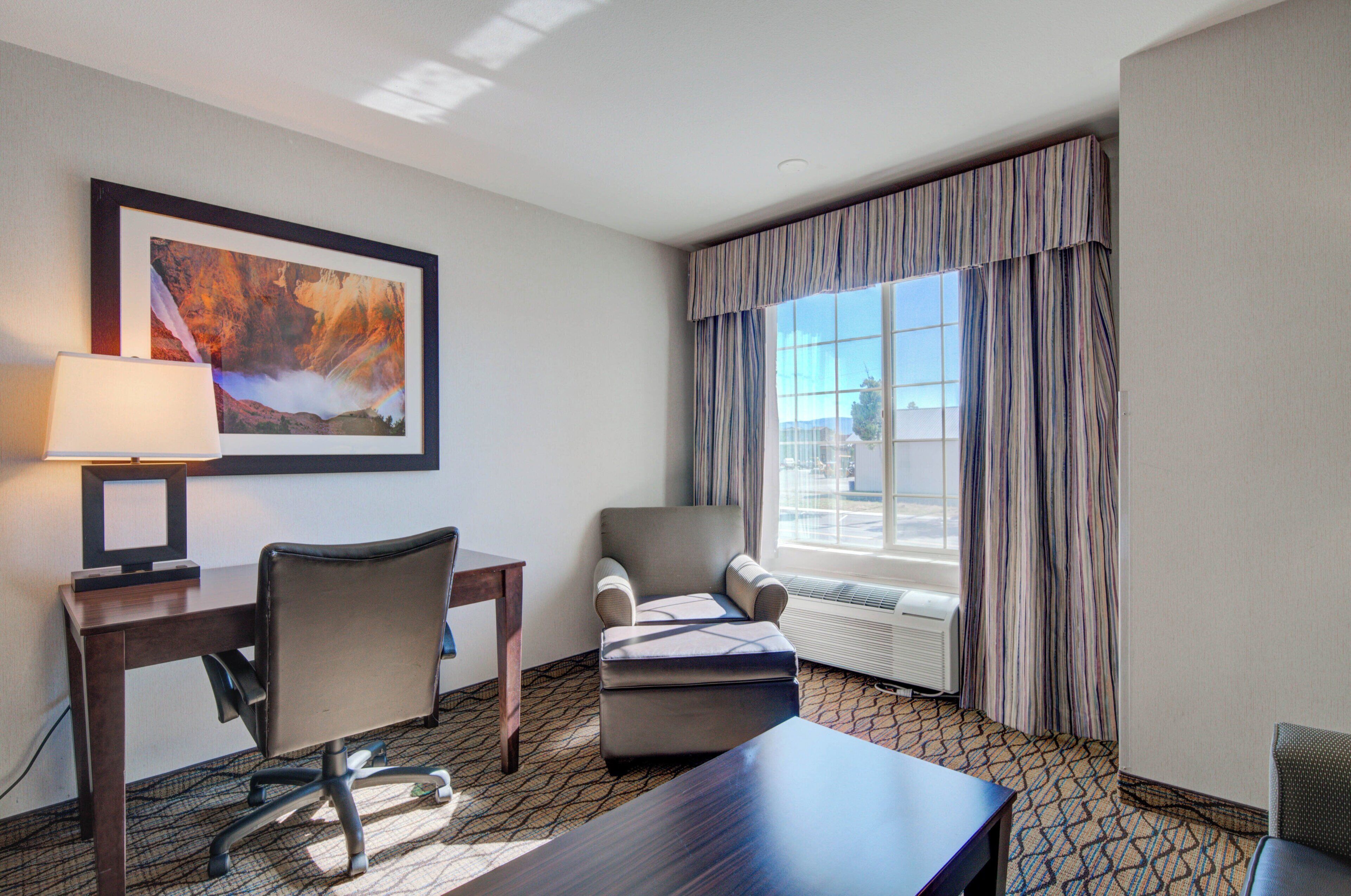 Une chambre d  ’hôtel équipée d’un bureau, d’une chaise, d’un pouf et d’une grande fenêtre avec des rideaux.