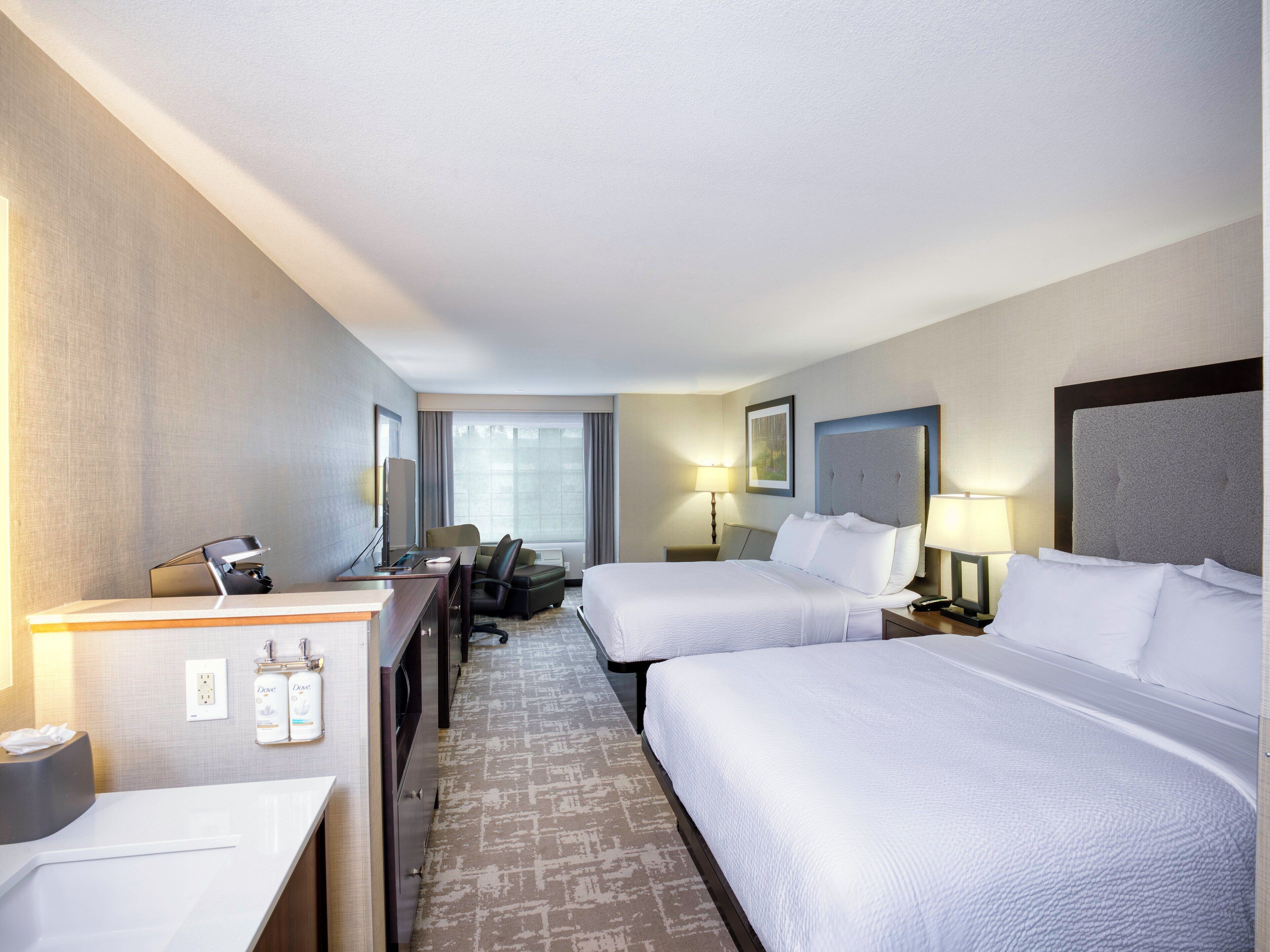 A hotel room with two beds, a desk, a chair, a lamp, and a window with curtains.
