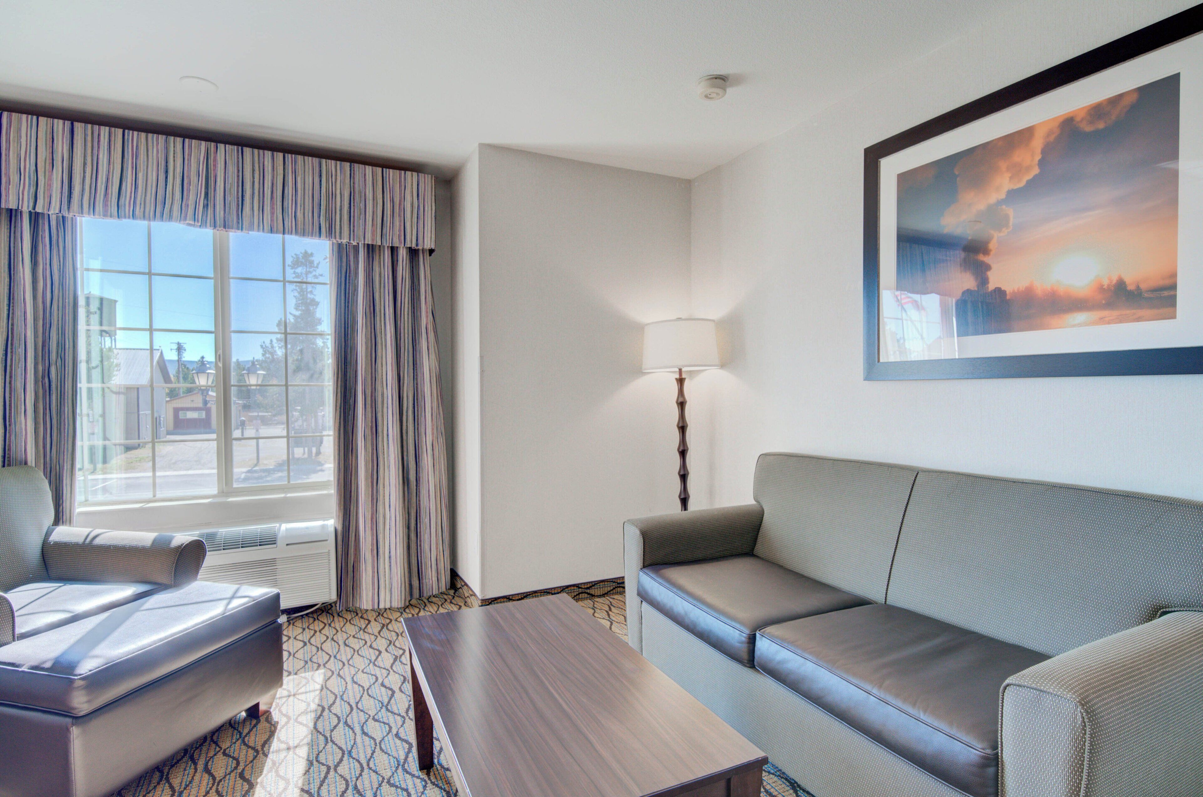 Une chambre d’hôtel avec une grande fenêtre, un canapé, un fauteuil, une lampe et un tableau au mur.