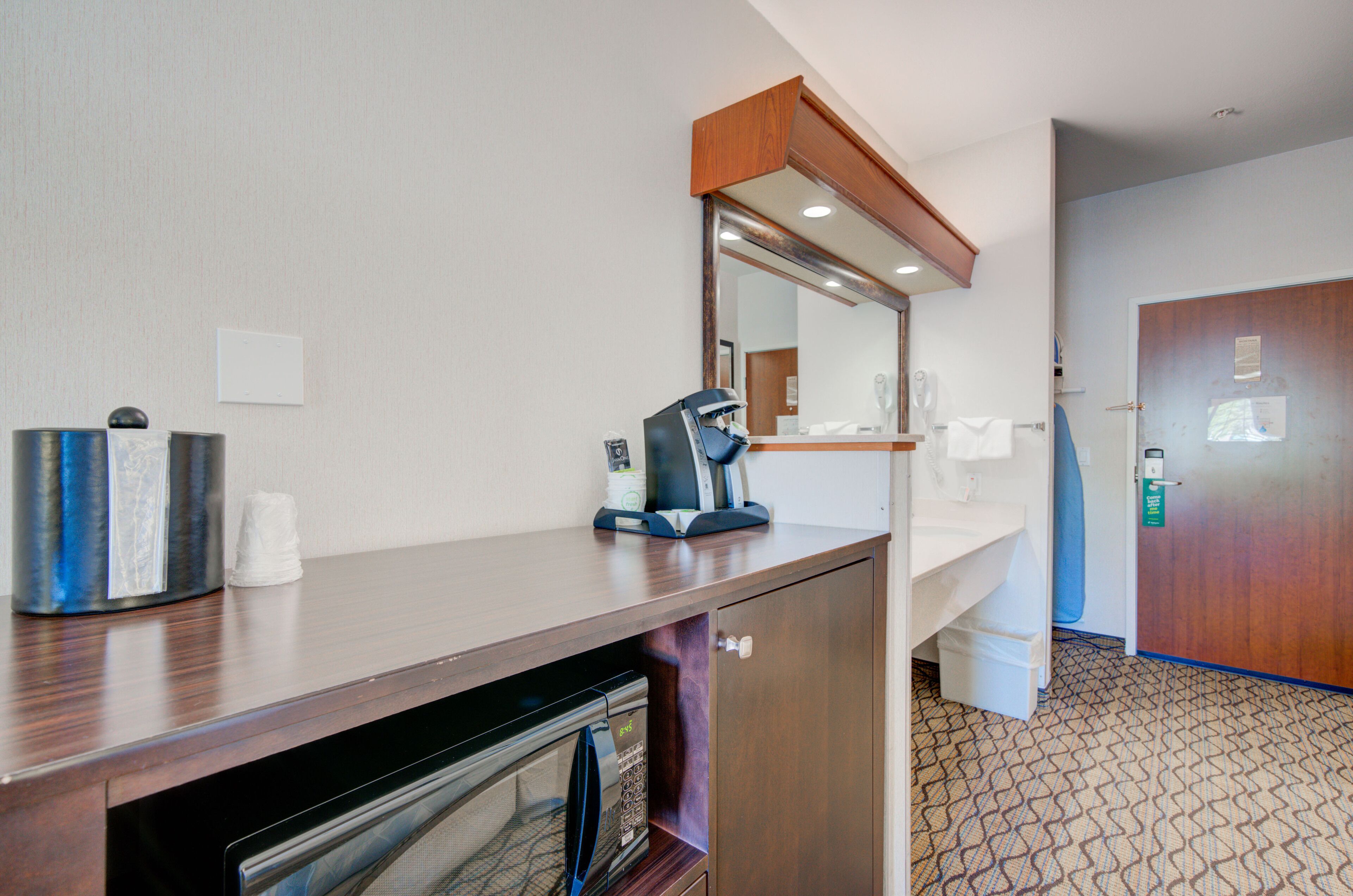 A hotel room with a wooden desk, a microwave, a coffee maker, and a door with a keycard lock.