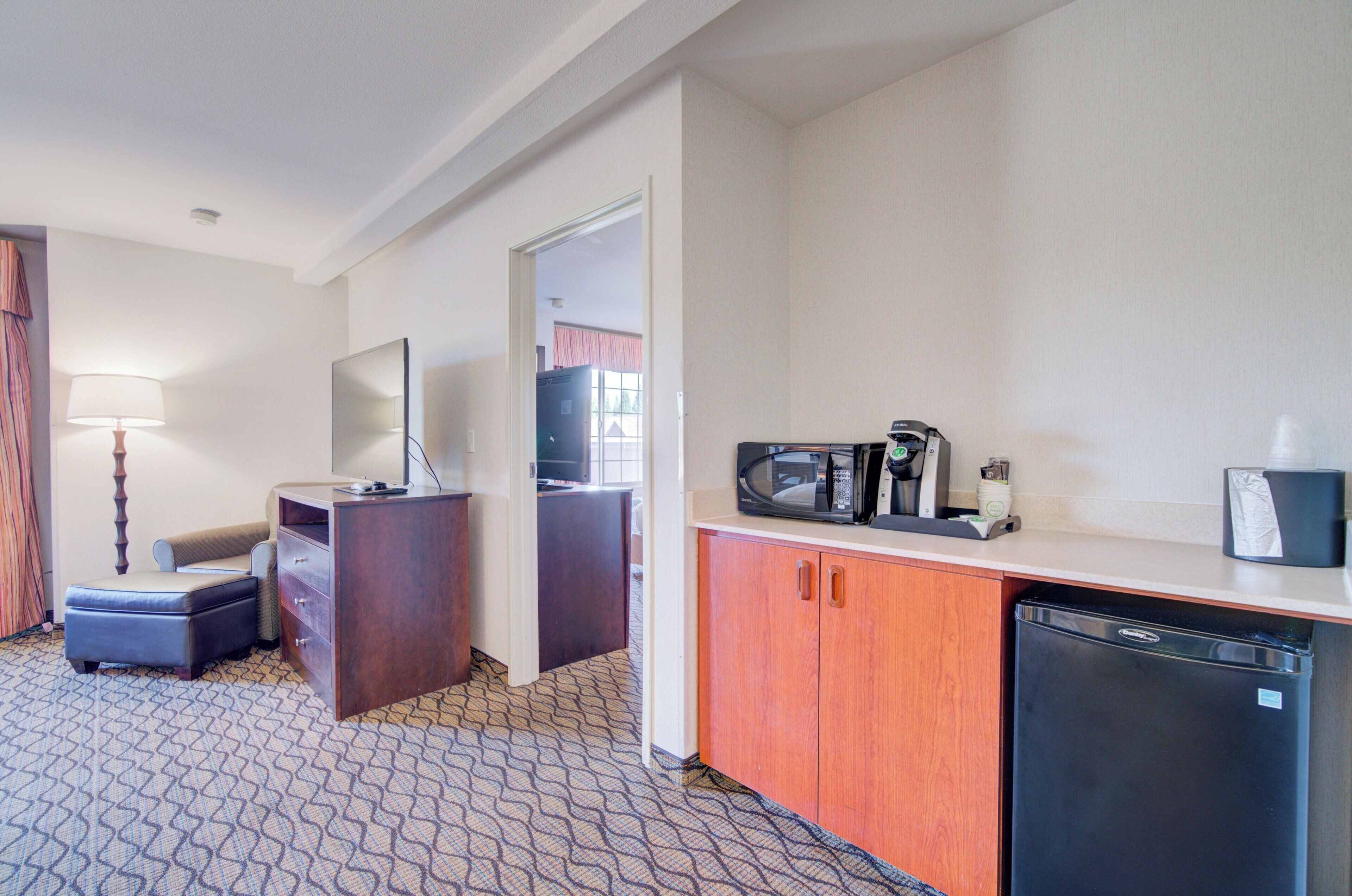 A compact hotel room with a kitchenette, a desk with a lamp, a chair, and a small seating area.