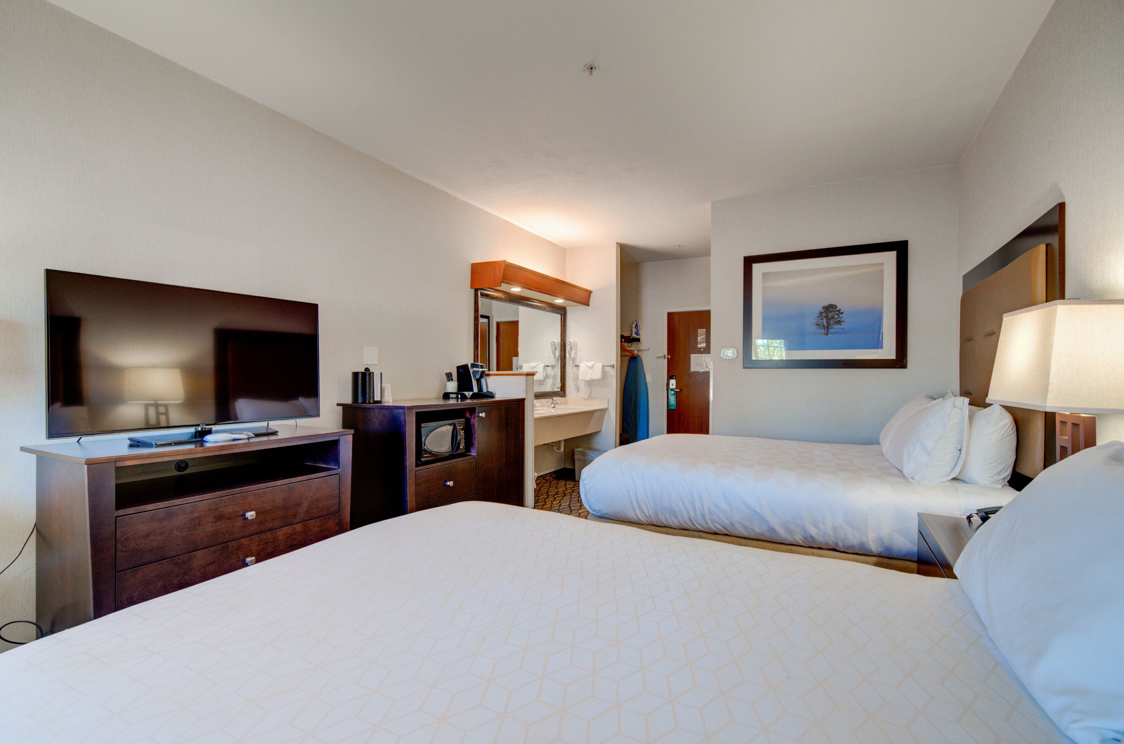 Habitación de hotel con dos camas, un televisor, un escritorio con computadora, un espejo y un cuadro en la pared.