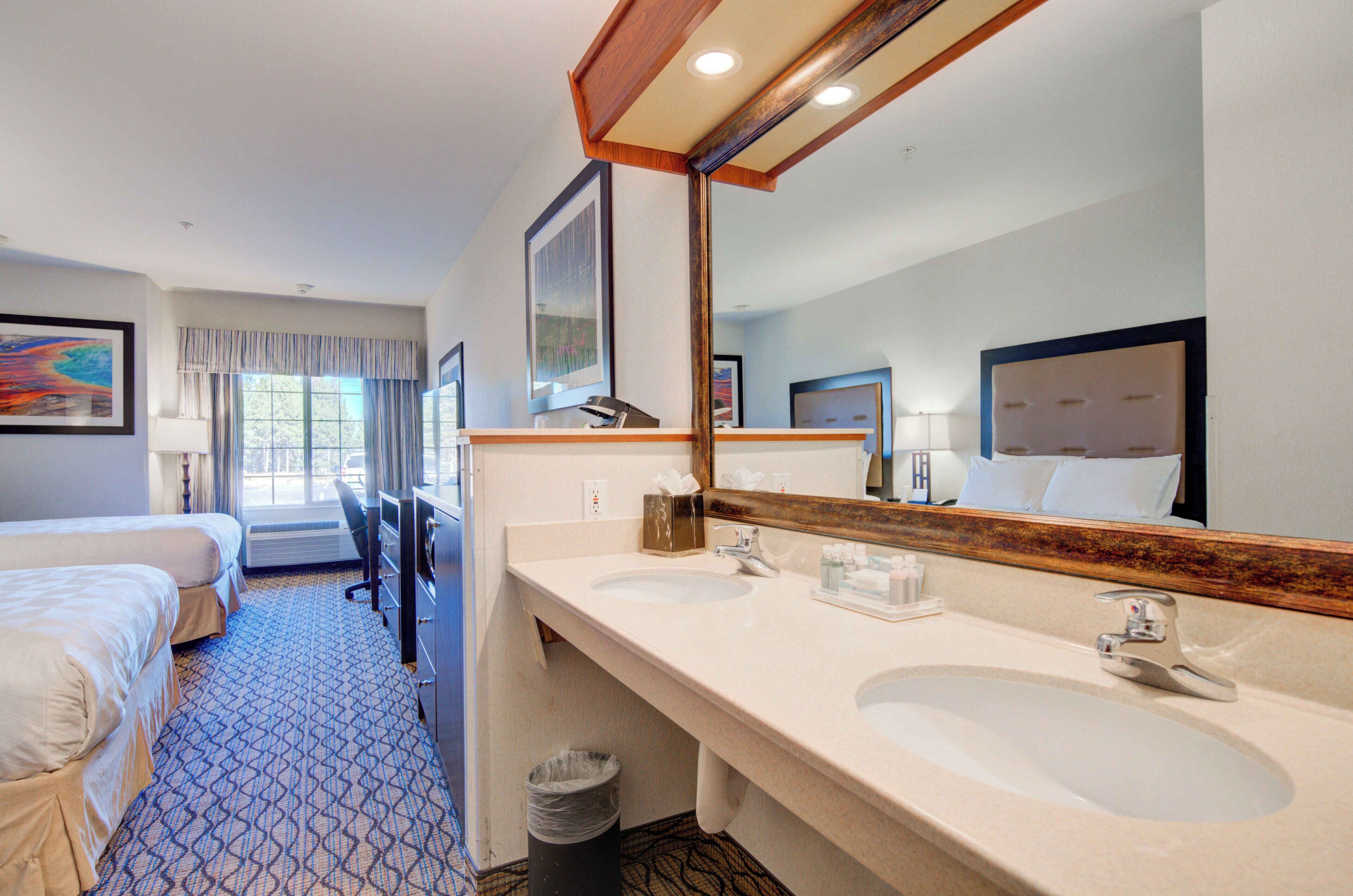 A hotel room with two beds, a large mirror, two sinks, and a trash can.