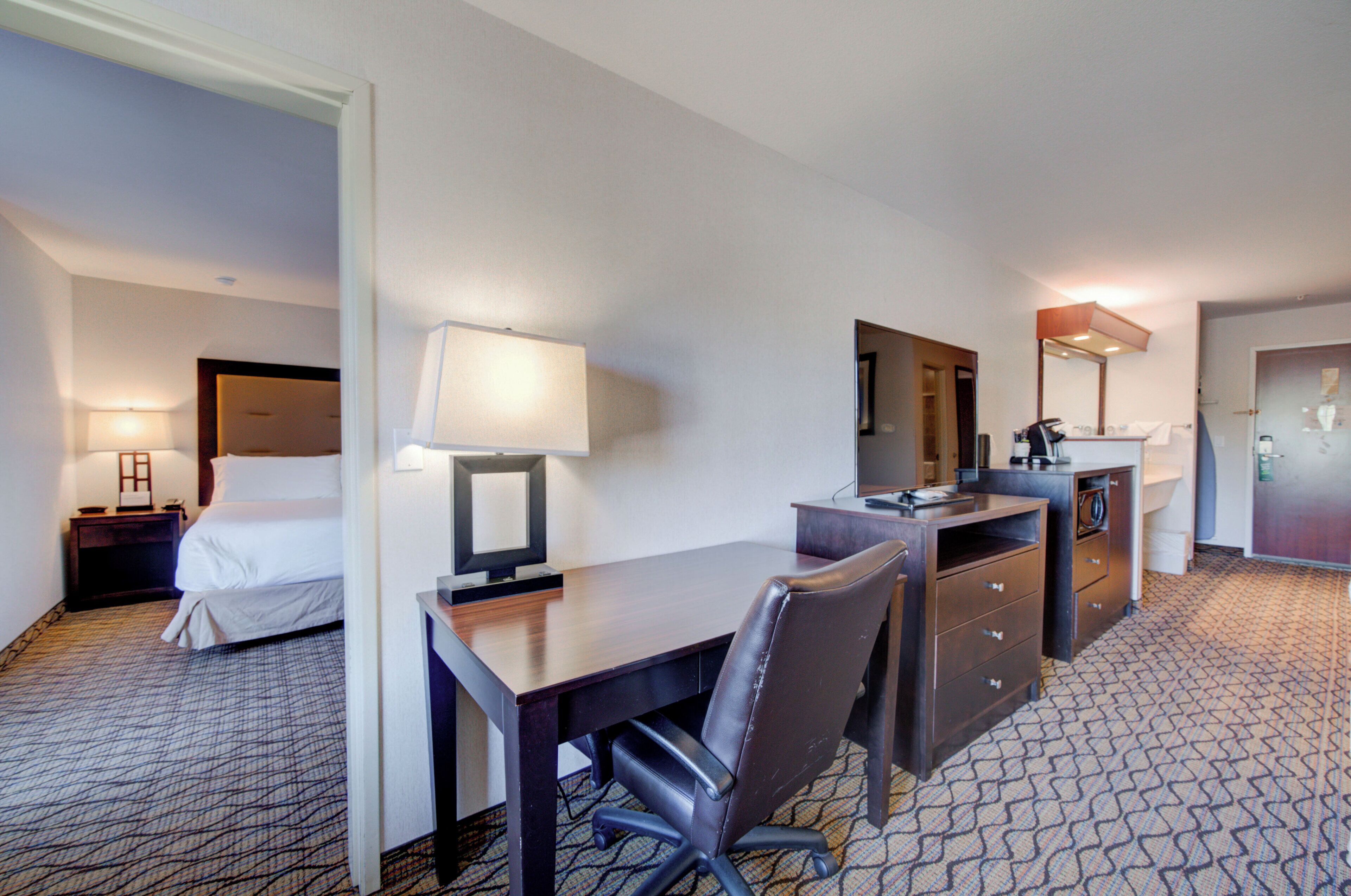 A hotel room with a bed, desk, chair, lamp, and a small kitchenette.