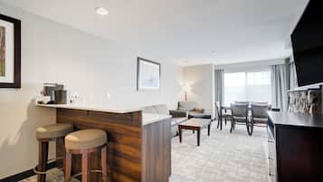 A hotel room with a kitchenette, a seating area with a sofa and chairs, a small table, and a bar area.