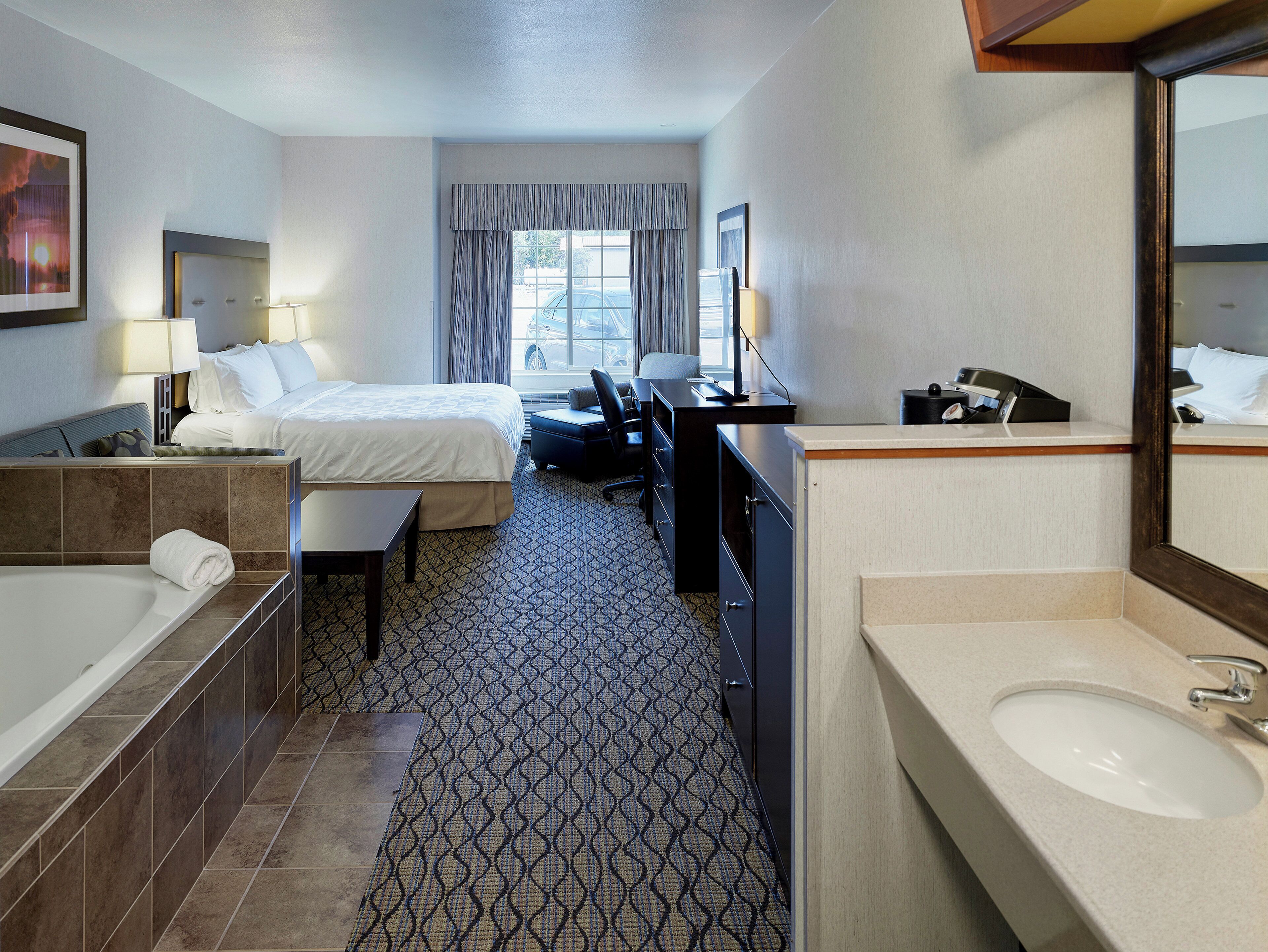 A hotel room with a bed, a desk, a chair, a desk lamp, a telephone, a bathroom with a sink and mirror, and a window with curtains.