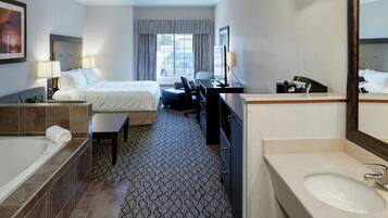 A hotel room with a bed, a desk, a chair, a desk lamp, a telephone, a bathroom with a sink and mirror, and a window with curtains.