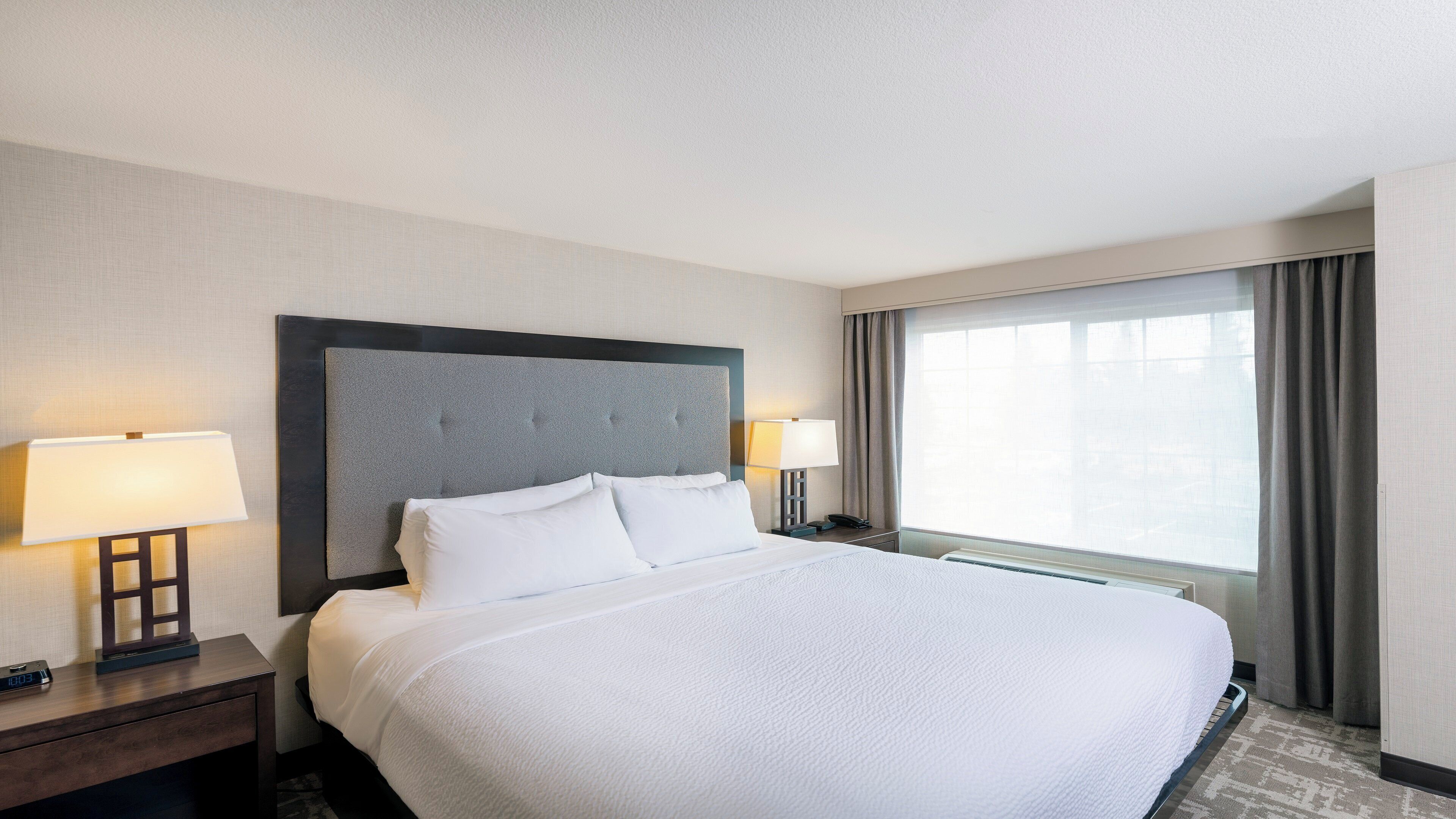 Une chambre d’hôtel avec un grand lit, deux tables de chevet avec des lampes, une fenêtre avec des rideaux et une horloge fixée au mur.