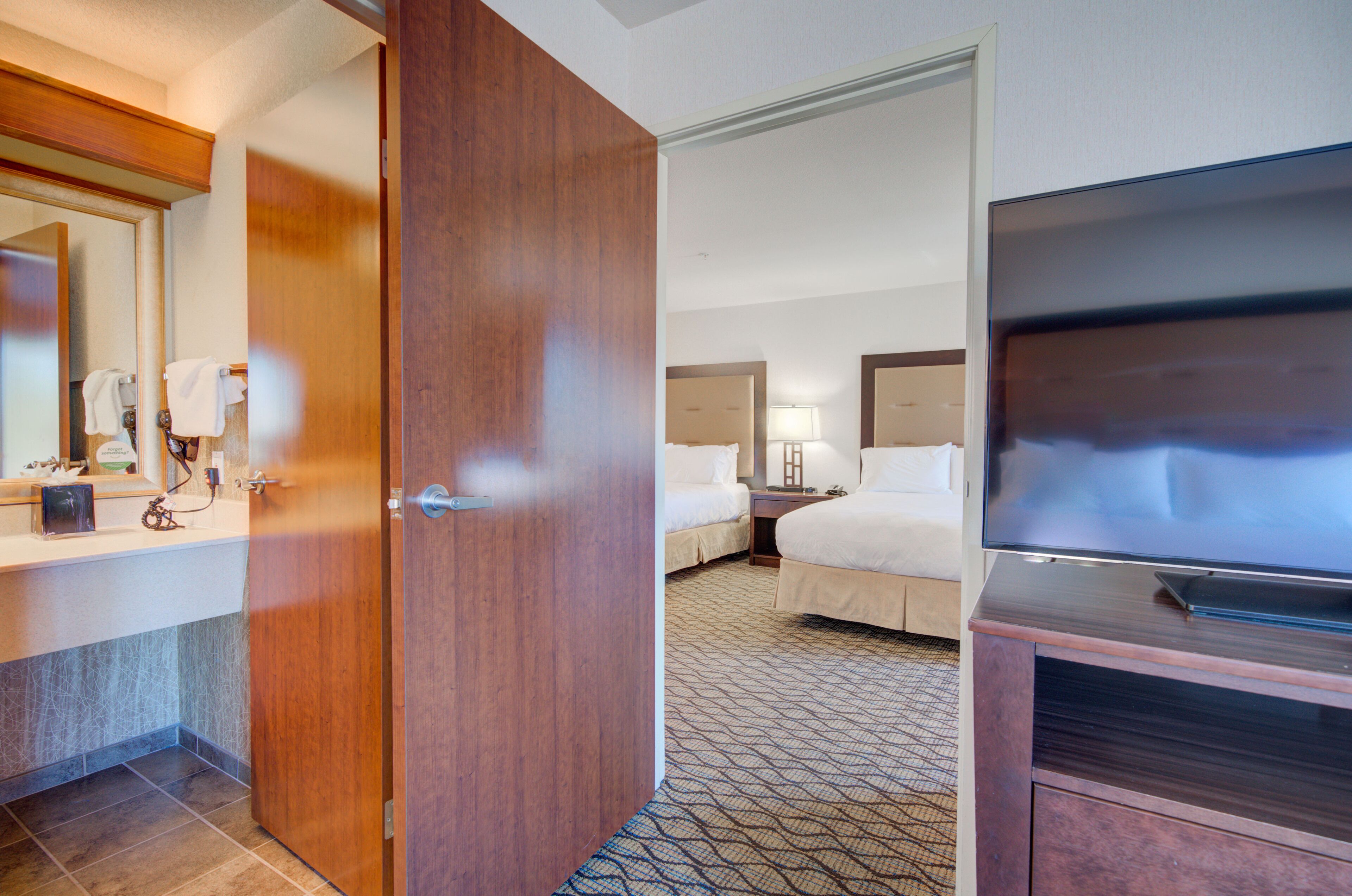 Habitación de hotel con puerta de madera, un espejo grande, un televisor de pantalla plana, una cama con cabecero y una alfombra estampada.