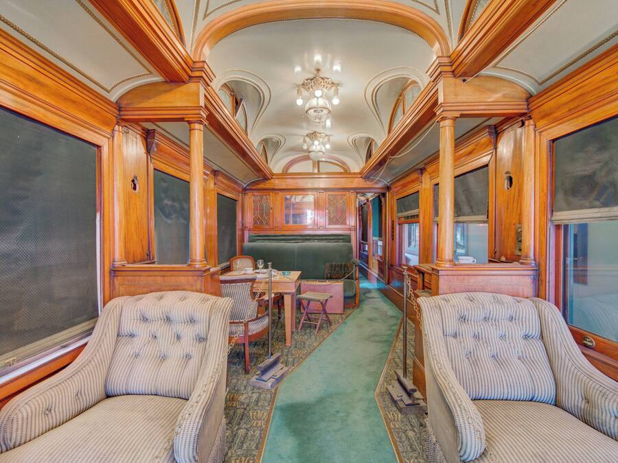 A train car interior with a green carpet, wooden paneling, upholstered chairs, a table, and a chandelier.