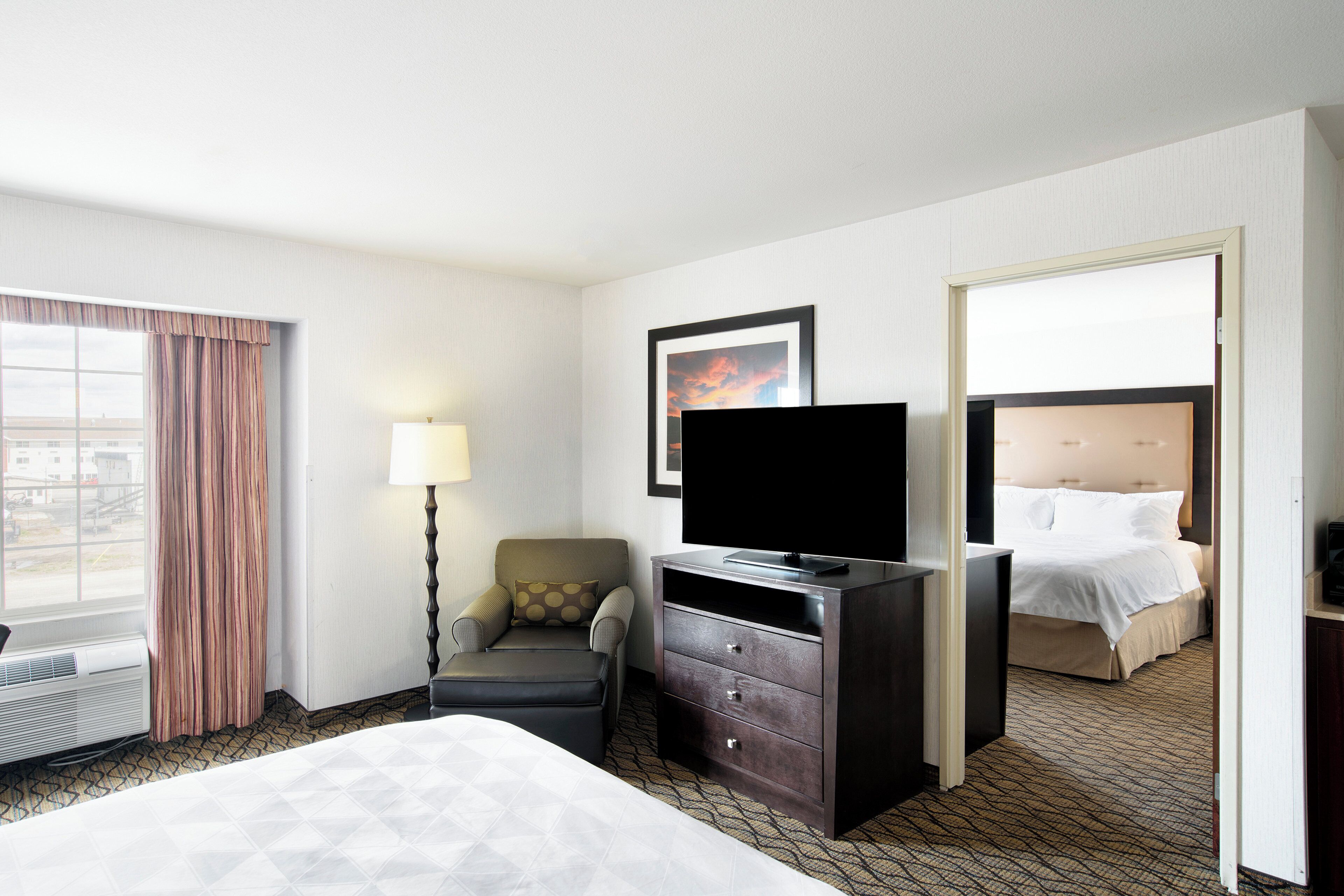 A hotel room with a bed, a desk with a flat-screen TV, a chair, a lamp, and a window with curtains.