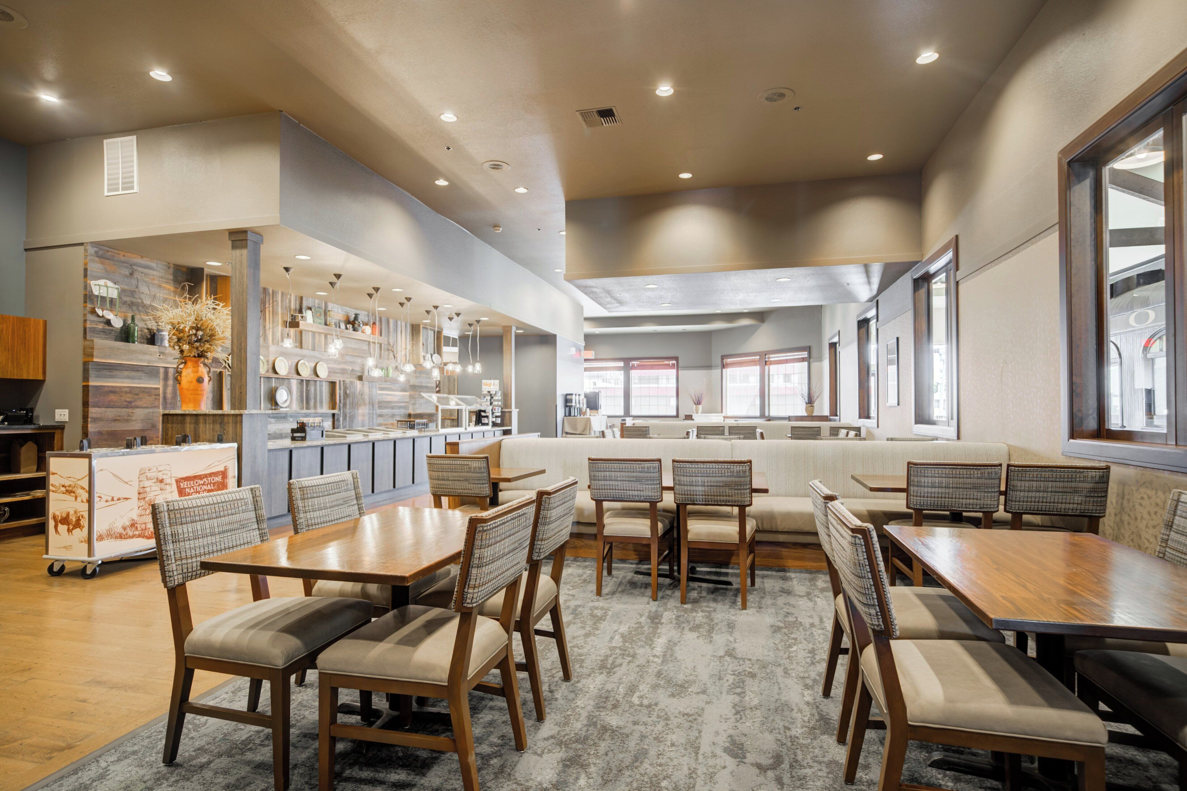 A spacious dining area with wooden tables and chairs, a kitchen in the background, and large windows providing natural light.