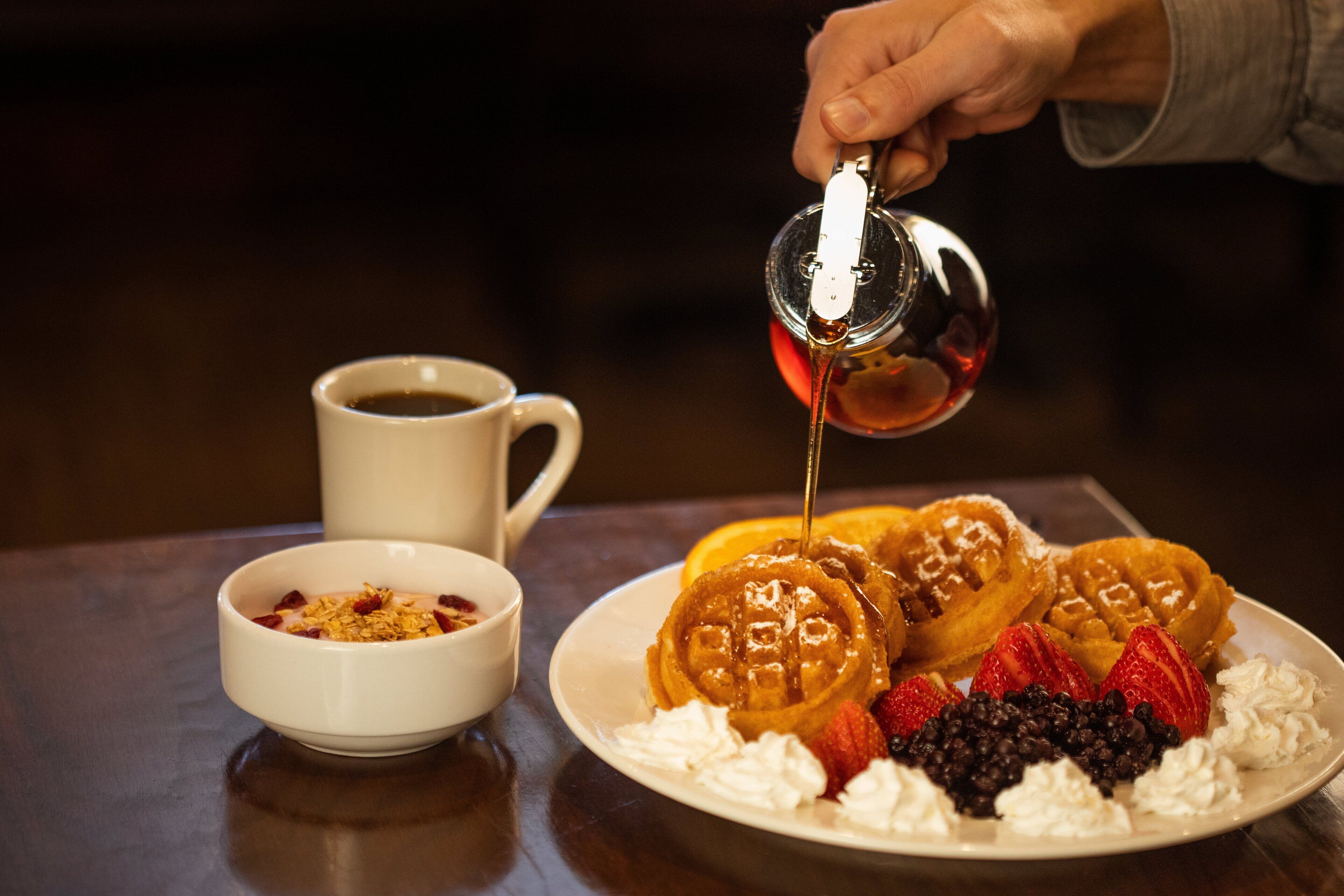 A plate of waffles with whipped cream and strawberries, a cup of coffee, and a bowl of cereal.