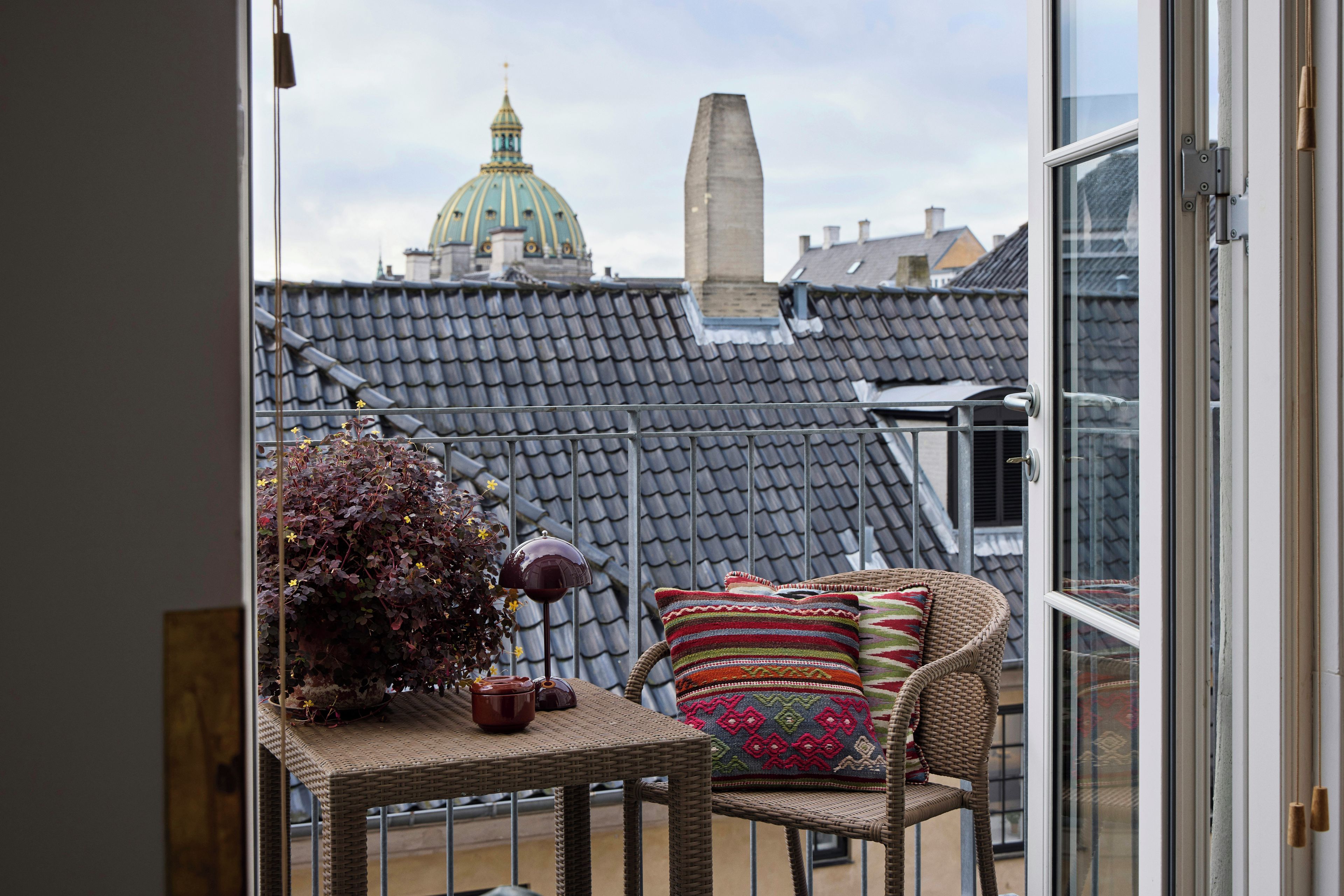 A balcony with a wicker chair, a small table with a vase and a teapot, and a view of a building with a dome and a chimney.