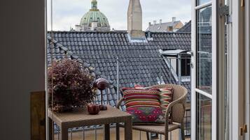 A balcony with a wicker chair, a small table with a vase and a teapot, and a view of a building with a dome and a chimney.