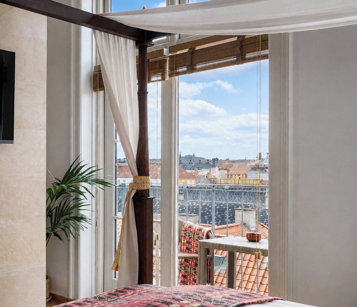 Una habitación con una cama, un ventanal con cortinas y vistas a tejados, además de una planta en maceta.