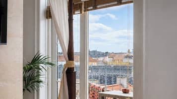 Una habitación con una cama, un ventanal con cortinas y vistas a tejados, además de una planta en maceta.
