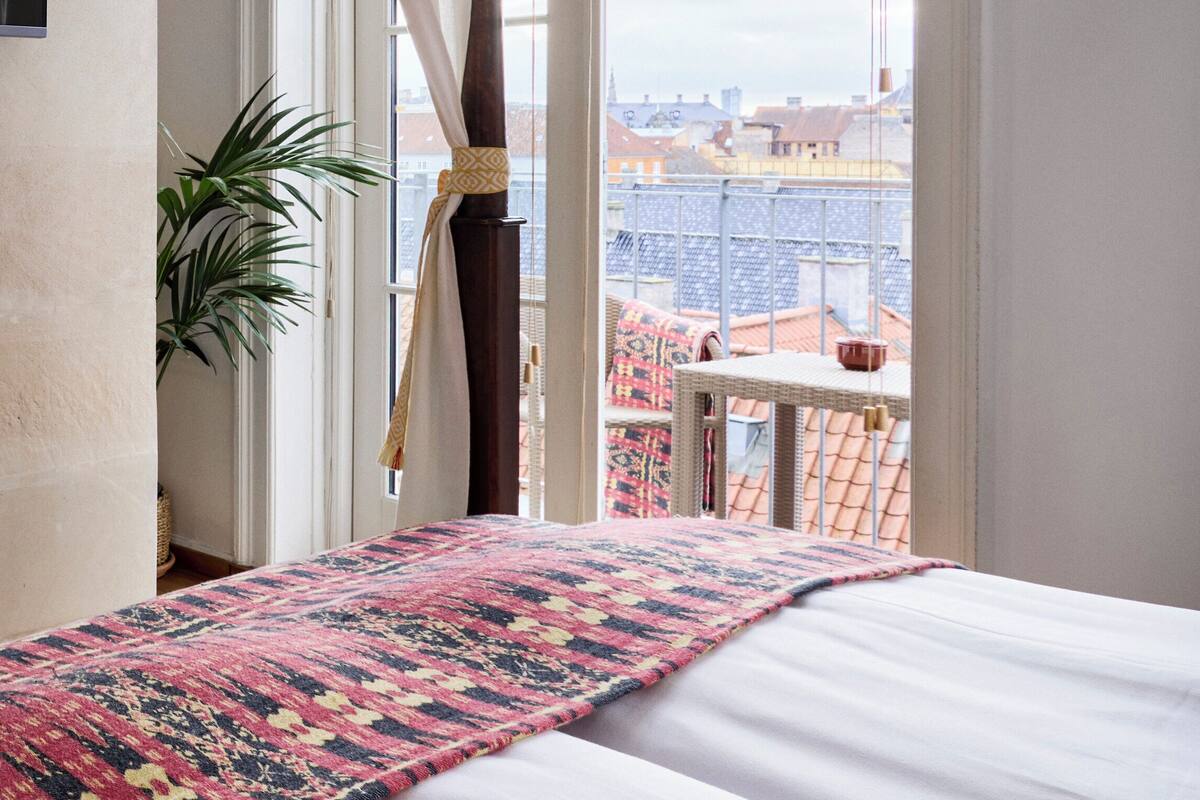 Una cama con una colcha estampada, una planta en maceta y vistas de un paisaje urbano a través de una ventana con persianas.