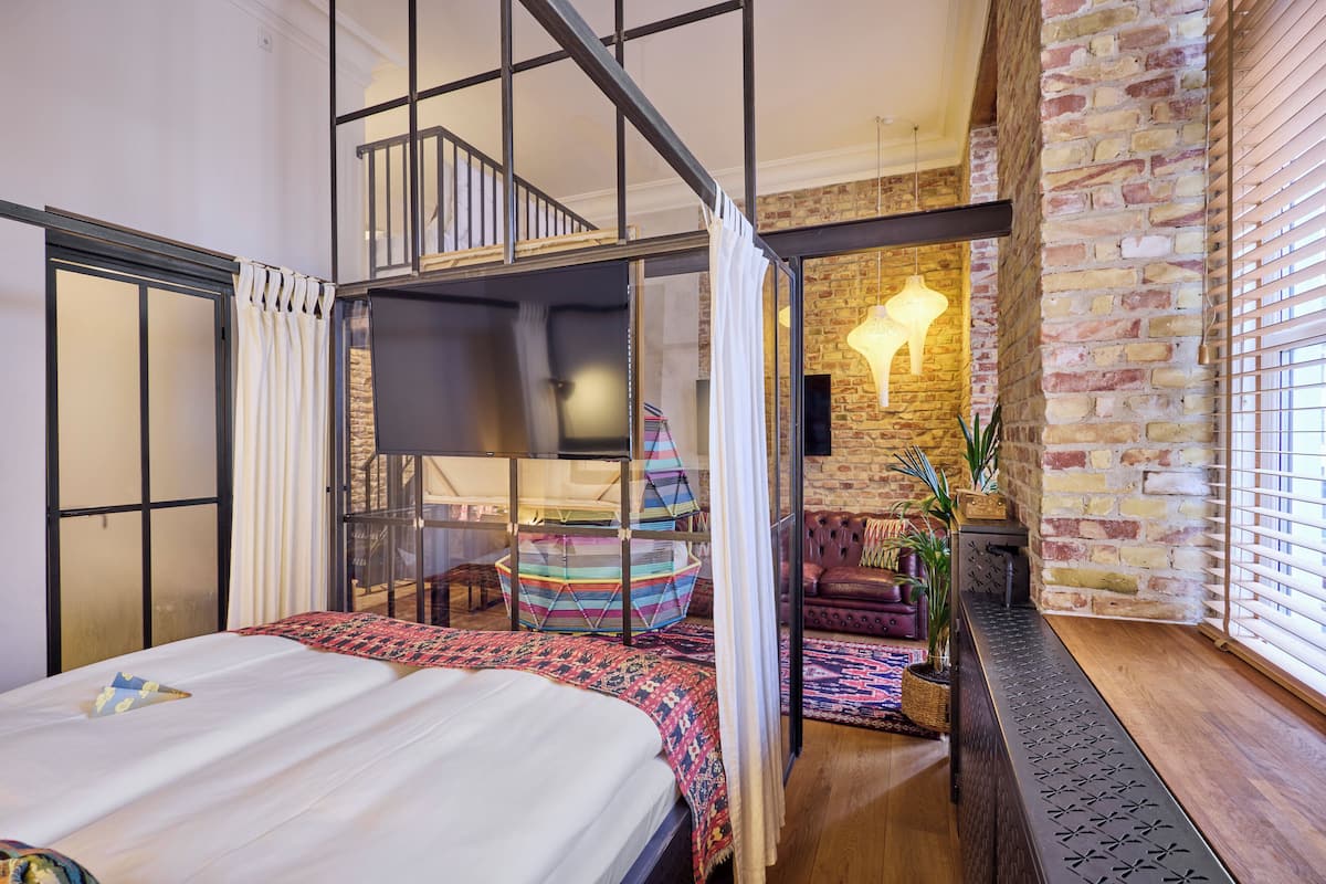 A modern bedroom with a large bed, a glass partition, a colorful hanging chair, and a brick wall.