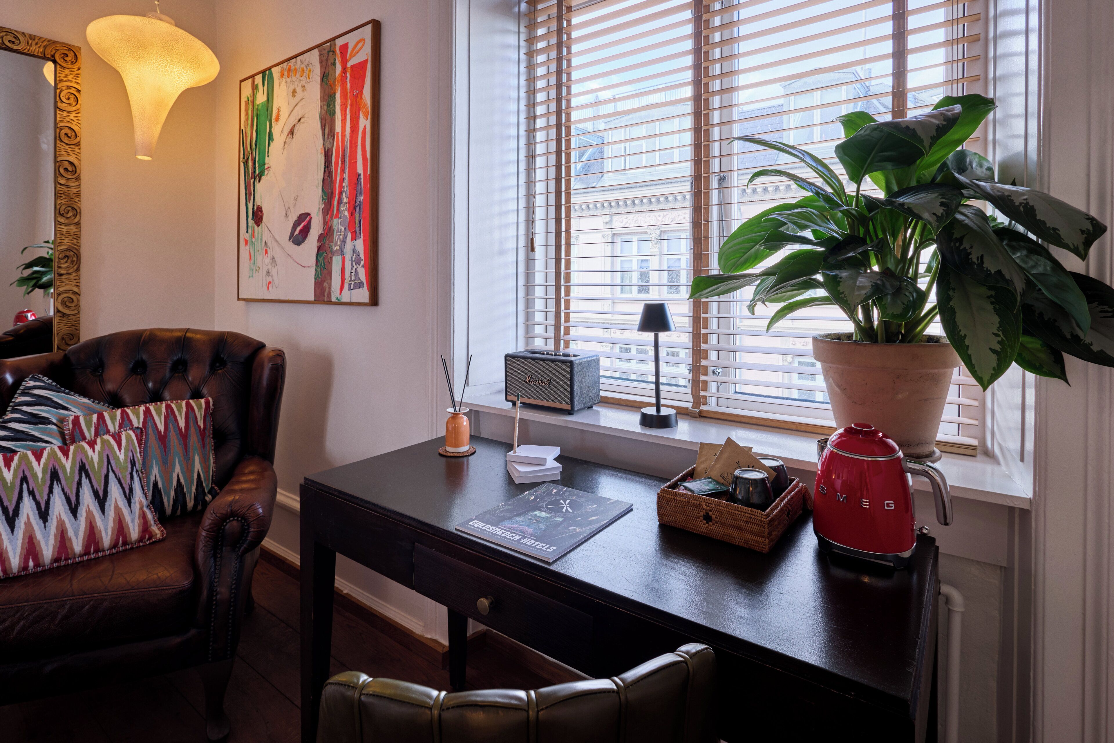 A modern living room with a leather armchair, a desk with a laptop, a potted plant, and a red kettle.