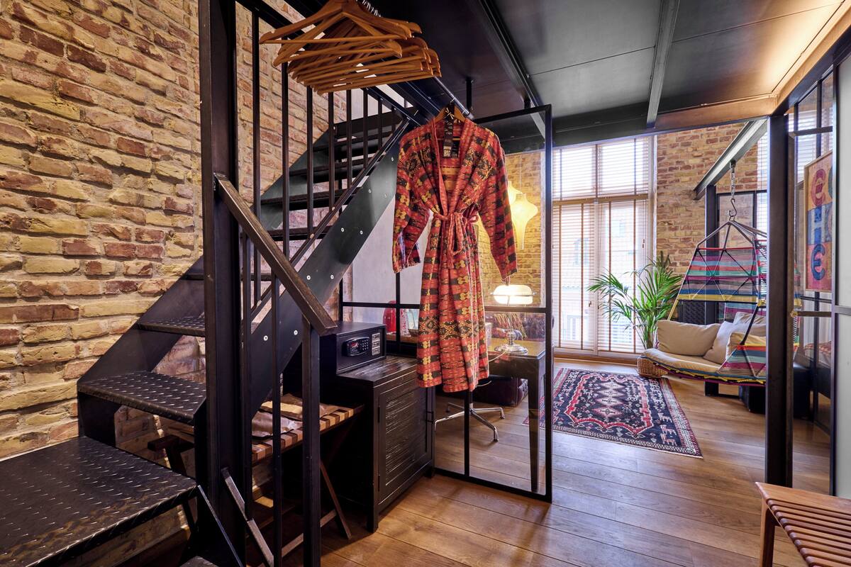 Una habitación acogedora con pared de ladrillo, una escalera de madera, una bata colgada, un escritorio y una zona de estar con cojines y una alfombra colorida.