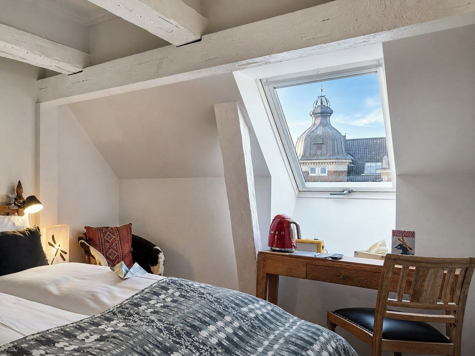 Un dormitorio con techo inclinado, cama con ropa de cama estampada, escritorio de madera, una silla y vista a un edificio a través de un tragaluz.