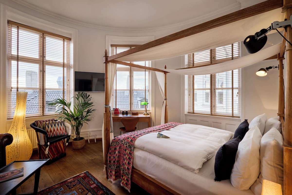 A bedroom with a four-poster bed, a desk with a chair, a television, and large windows with blinds.