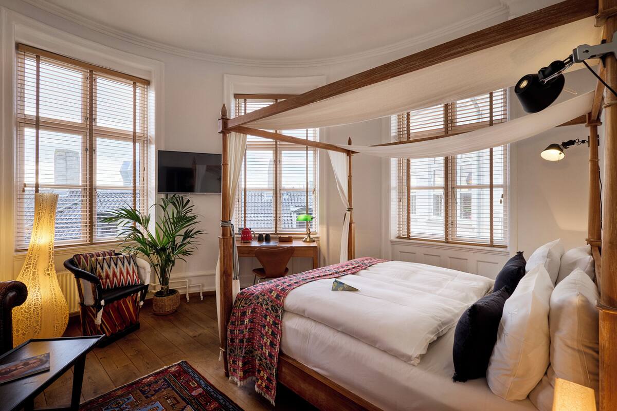 Un dormitorio con cama con dosel, un escritorio con silla, televisor y ventanas grandes con persianas.
