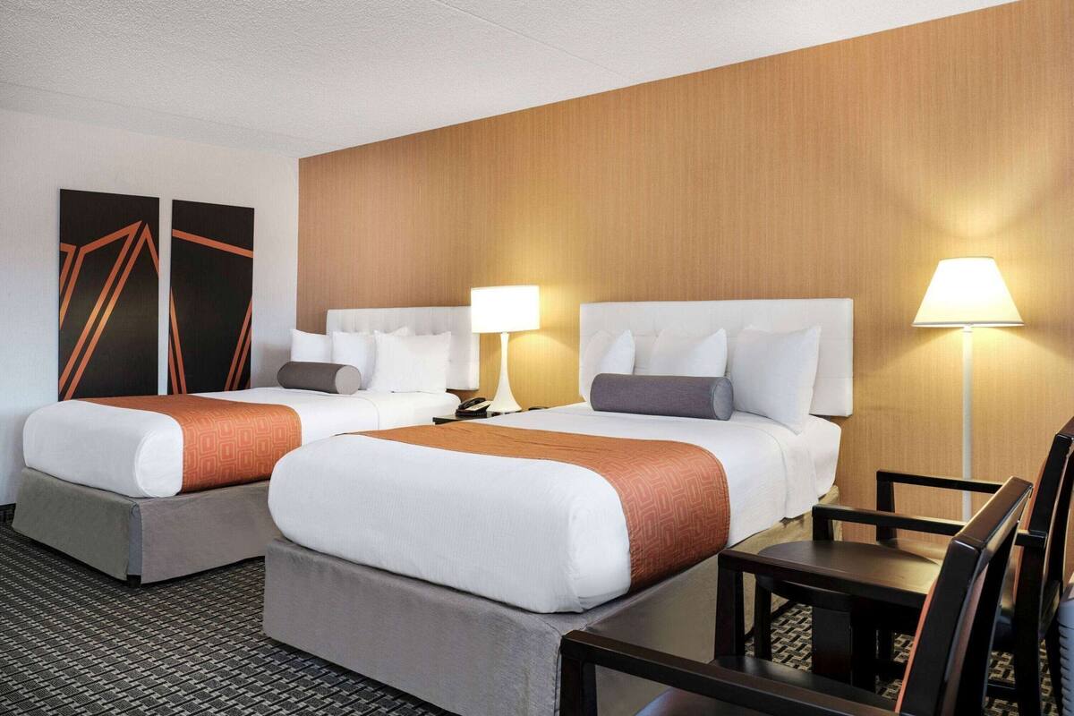 A hotel room with two beds, a desk, a chair, and a lamp.