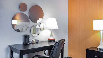 A hotel room with a desk, a chair, a coffee maker, a lamp, and a mirror.