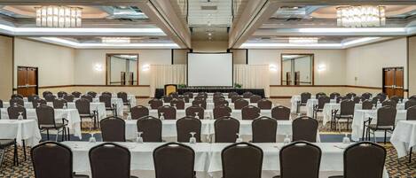 A conference room with rows of tables and chairs, a large screen at the front, and chandeliers on the ceiling.