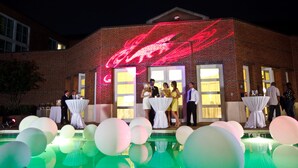 A brick building with a red neon sign, a pool with white and green balloons, and tables with white tablecloths.