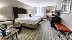 A hotel room with two beds, a desk, a chair, a bench, and a small table with a lamp.