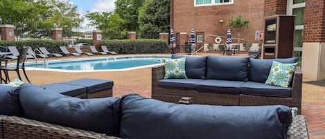 A poolside area with lounge chairs, a wicker sofa, and a brick building in the background.