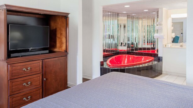 A hotel room with a large bed, a wooden dresser with a flat-screen TV, and a red jacuzzi tub. - Howard Johnson by Wyndham Niagara Falls (Niagara Falls)