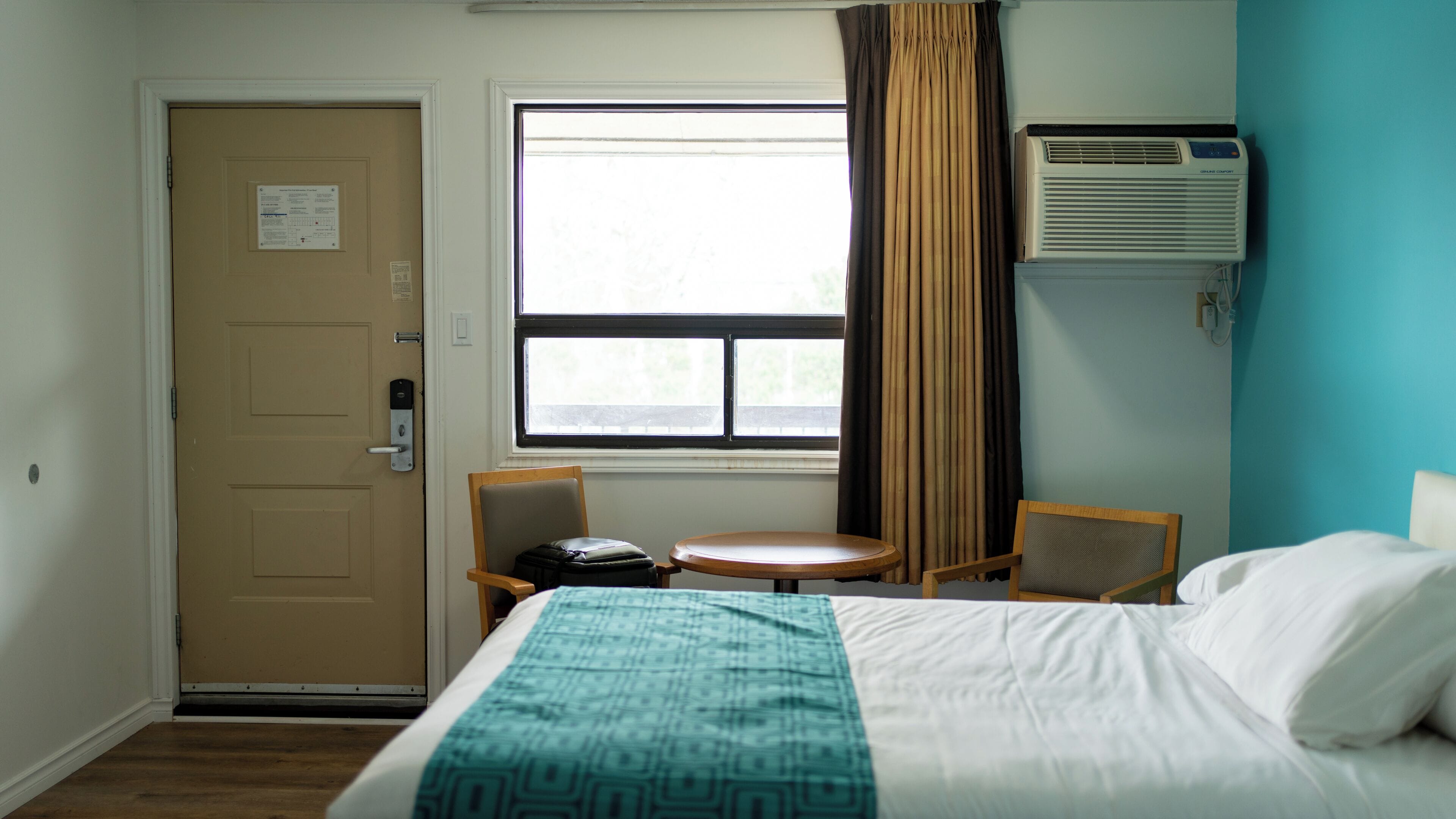 A hotel room with a bed, a desk, a chair, a window, an air conditioning unit, and a door.