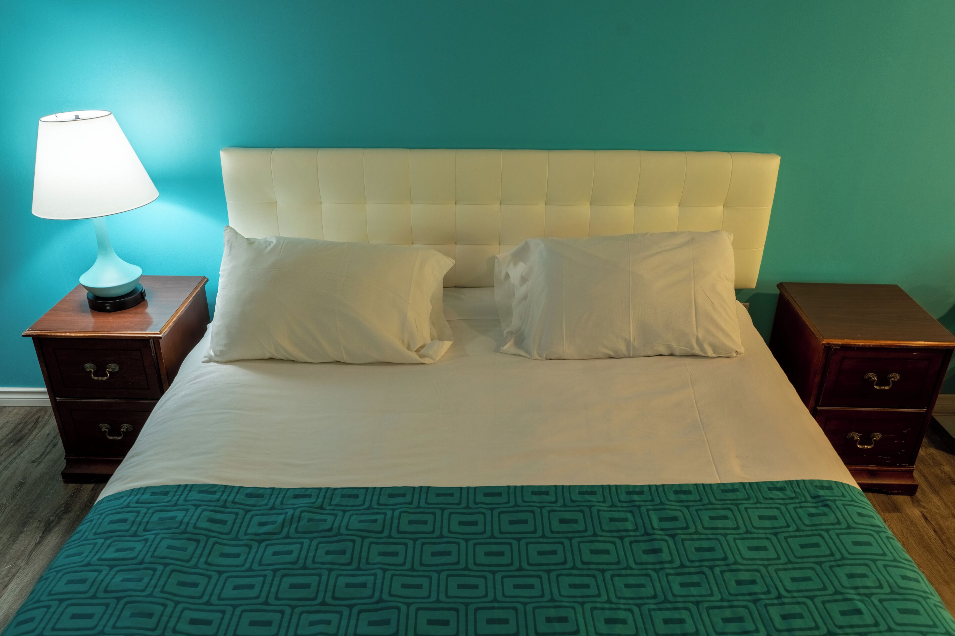 A bed with a white headboard, two pillows, a green patterned bedspread, and a bedside lamp on a wooden nightstand.