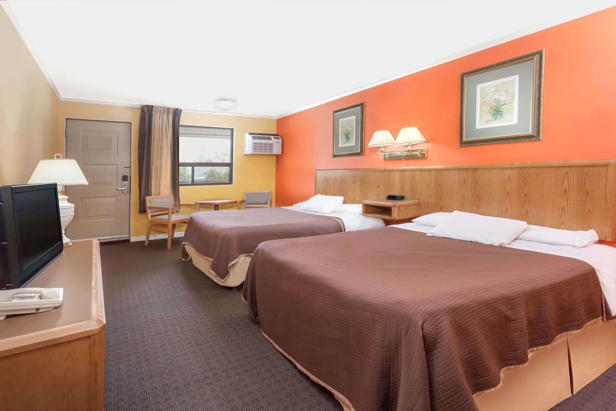 Ein Hotelzimmer mit zwei Betten, einem Schreibtisch mit Fernseher, einem Sessel, einem Fenster und einer Klimaanlage.