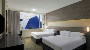 A hotel room with two beds, a TV, a desk, and a window with a view of a blue building.