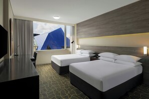 A hotel room with two beds, a TV, a desk, and a window with a view of a blue building.