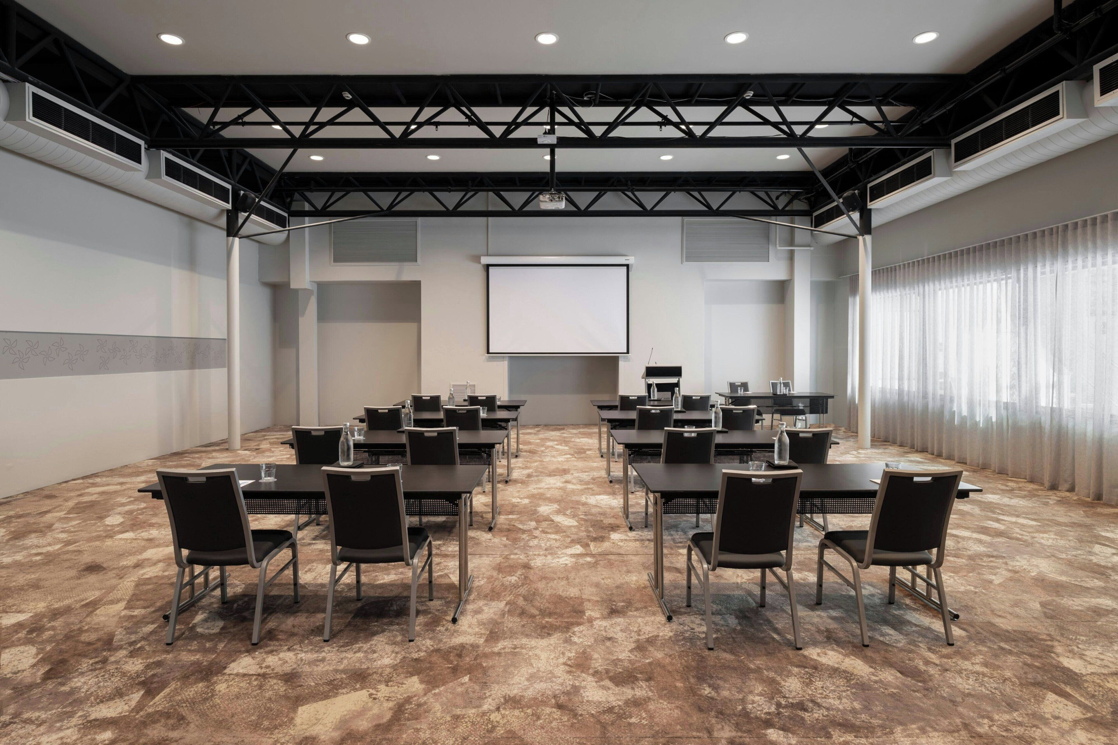 Un salón de conferencias con pantalla de proyector, varias mesas y sillas dispuestas para reuniones.