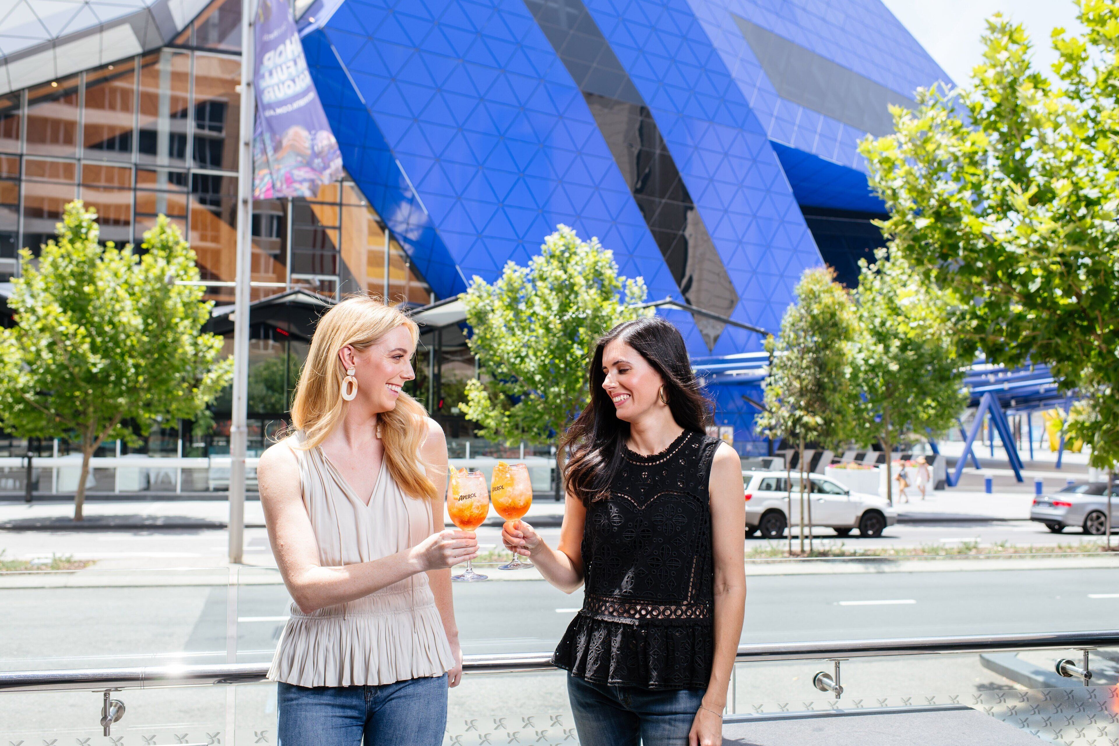 Two women toasting with drinks in front of a modern building with a blue glass facade.